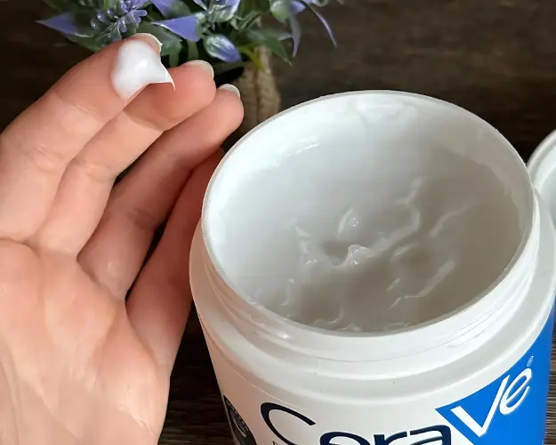 Hand holding a small amount of white cream scooped out from a jar of CeraVe cream